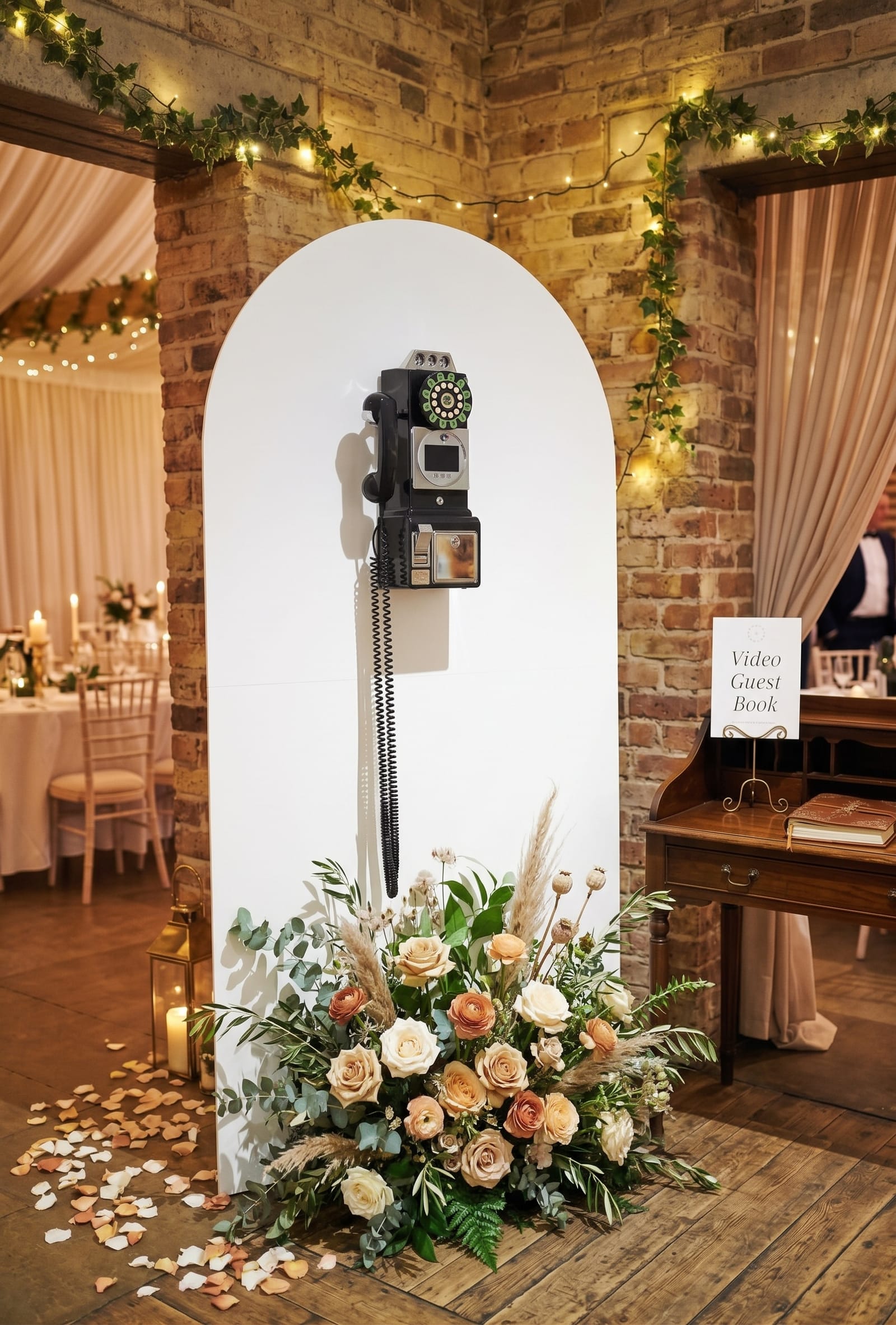 A vintage black rotary payphone video guestbook mounted on a tall white arched panel at a candlelit wedding venue with exposed brick walls, ivy garlands and twinkling fairy lights. Its coiled cord hangs above a lush floor arrangement of peach and ivory roses, ranunculus, pampas grass and eucalyptus, surrounded by scattered petals and a brass lantern. An antique writing desk beside it displays a 'Video Guest Book' sign, with a softly glowing reception room of draped ceilings and set tables beyond.